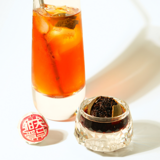 Glass of iced tea with a orange tea, a glass teacup with loose tea leaves, and a tea ball on a white background.