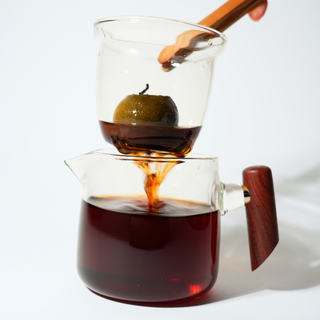 Glass pitcher with wooden handle and handle pouring tea into another glass container.