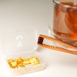 Gold tea resins  in a small white container with wooden tongs and a glass of tea in the background.