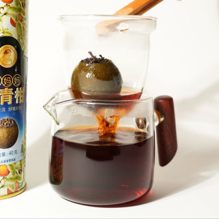 Tea packaging with a glass teapot containing a green tea ball and a wooden spoon.