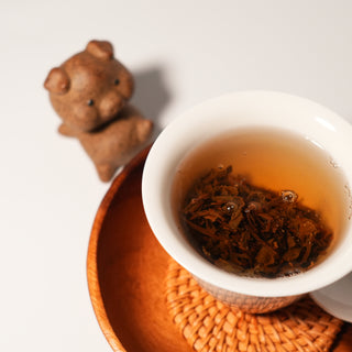 The whole tea leaves from a Yustea mini Pu'er cake, fully unfurled after brewing in a white cup, with a traditional bear tea pet in the background.