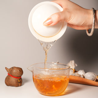 A traditional Gongfu tea ceremony in progress, showing tea being poured from a white gaiwan into a glass fairness pitcher, with a tea pet and Pu'er Dragon Balls nearby.
