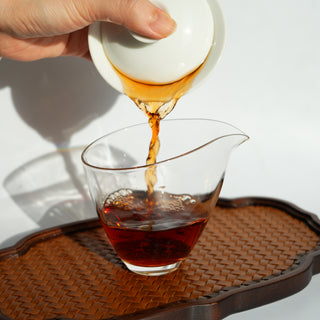  person performing a Gongfu tea ceremony, pouring dark, rich tea from a white porcelain gaiwan into a glass fairness pitcher (gong dao bei)