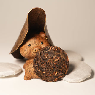 A whimsical photo of a piggy tea pet wearing a leaf hat, presenting a Yustea mini Pu'er tea cake with golden tips.