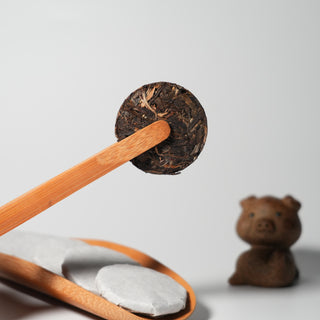 A close-up of an unwrapped mini Pu'er tea cake with golden tips, held for inspection by bamboo tongs, with a pig tea pet in the background.