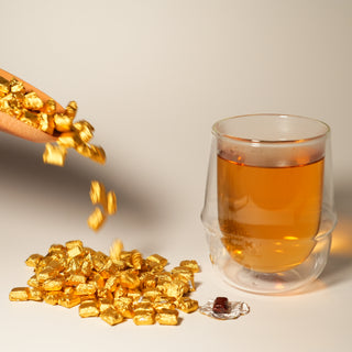 A dynamic shot of golden foil-wrapped tea resin tumbling from a wooden scoop, with a double-walled glass of brewed tea and an unwrapped piece of resin nearby.