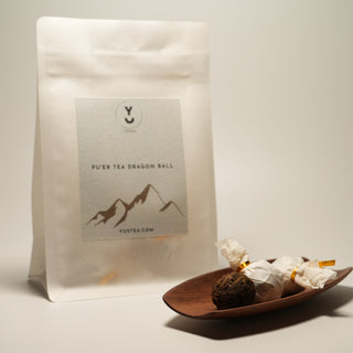 A product shot of Yustea's Pu'er Tea Dragon Ball, showing the retail pouch next to a wooden dish of wrapped and unwrapped tea balls.