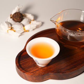 A traditional tea tasting setup, featuring a porcelain cup filled with golden Pu'er tea brewed from Yustea's Dragon Balls, with a serving pitcher and the tea balls in the background.