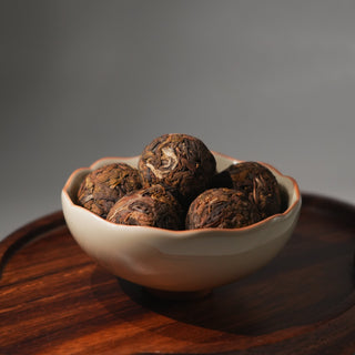 A ceramic bowl with a scalloped rim filled with hand-rolled Pu'er Dragon Balls, showcasing the compressed and aged whole tea leaves on a wooden tray.