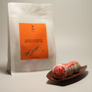 A product shot of Yustea's Green Mandarin Orange Pu'er Tea, showing the retail pouch next to three individually wrapped Xiaoqinggan portions in a wooden dish.