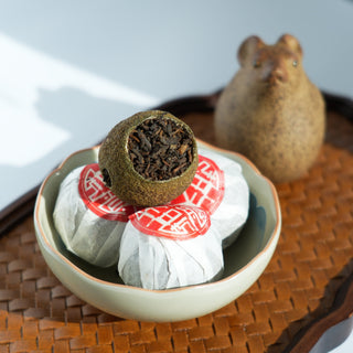 A bowl of Yustea's Xiaoqinggan Green Mandarin Pu'er, with one unwrapped to show the whole citrus peel filled with tea, and a traditional tea pet in the background