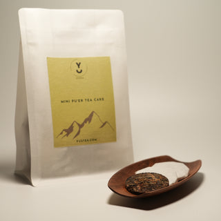 A product shot of Yustea's Mini Pu'er Tea Cake, showing the retail pouch next to a wooden dish containing one unwrapped and two wrapped tea cakes.