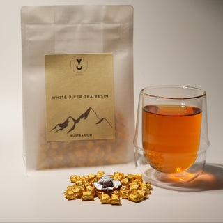 A product shot of Yustea's White Pu'er Tea Resin, showing the retail pouch, a pile of golden-wrapped resin, and a finished glass of brewed tea.